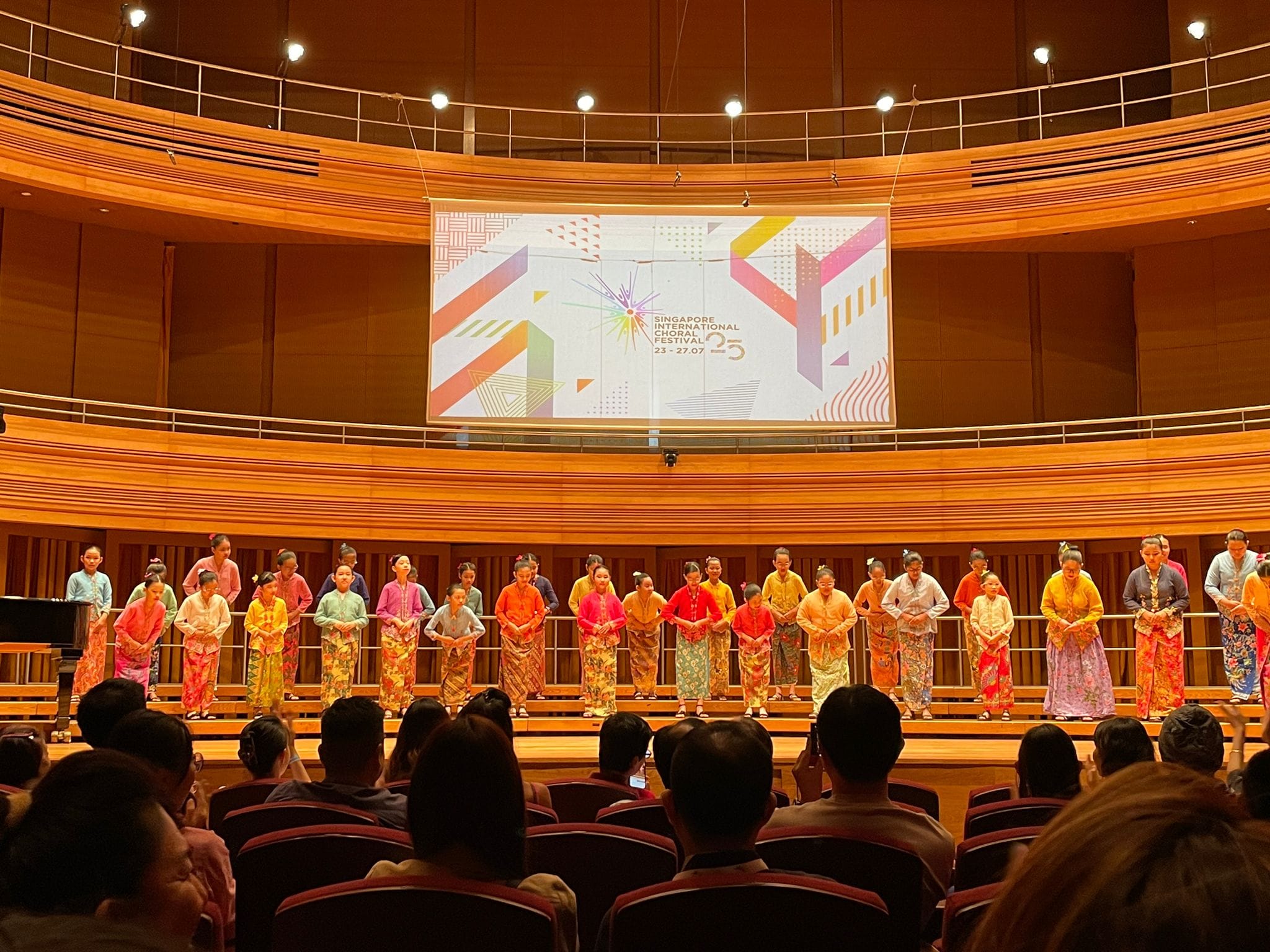 A choir in colorful traditional dress bows on stage at the Singapore International Choral Festival in a wood-paneled hall.